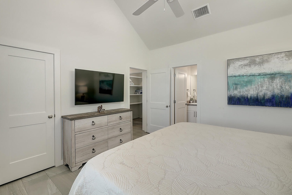 Your serene bedroom features coastal artwork, a mounted TV, and vaulted ceilings with a ceiling fan for ultimate comfort.