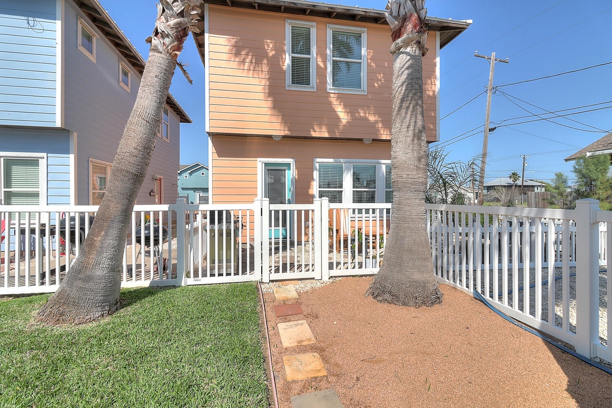 Two-story coastal home with orange siding and white trim, featuring a fenced front yard with palm trees in a residential neighborhood.