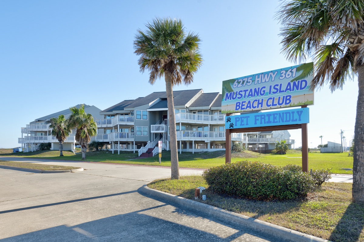 Mustang Island Beach Club welcomes visitors with its colorful entrance sign and coastal architecture surrounded by swaying palm trees.