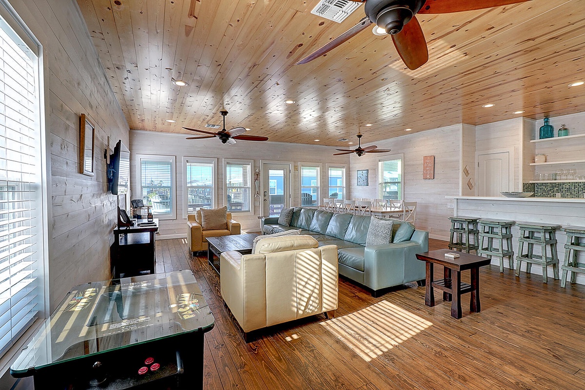 Relax in your spacious open-concept living area with soaring wood ceilings, coastal-inspired furnishings, and abundant natural light streaming through expansive windows.