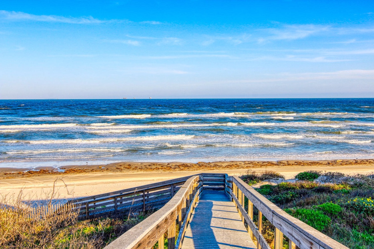 A wooden boardwalk leads through coastal dunes to pristine sandy shores and rolling ocean waves under brilliant blue skies.