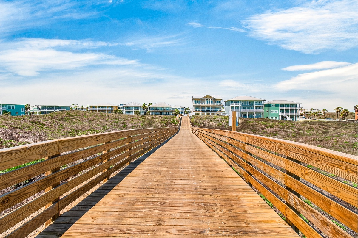 Wooden boardwalk leads through coastal dunes to beachfront properties with bright blue and turquoise facades under clear skies.