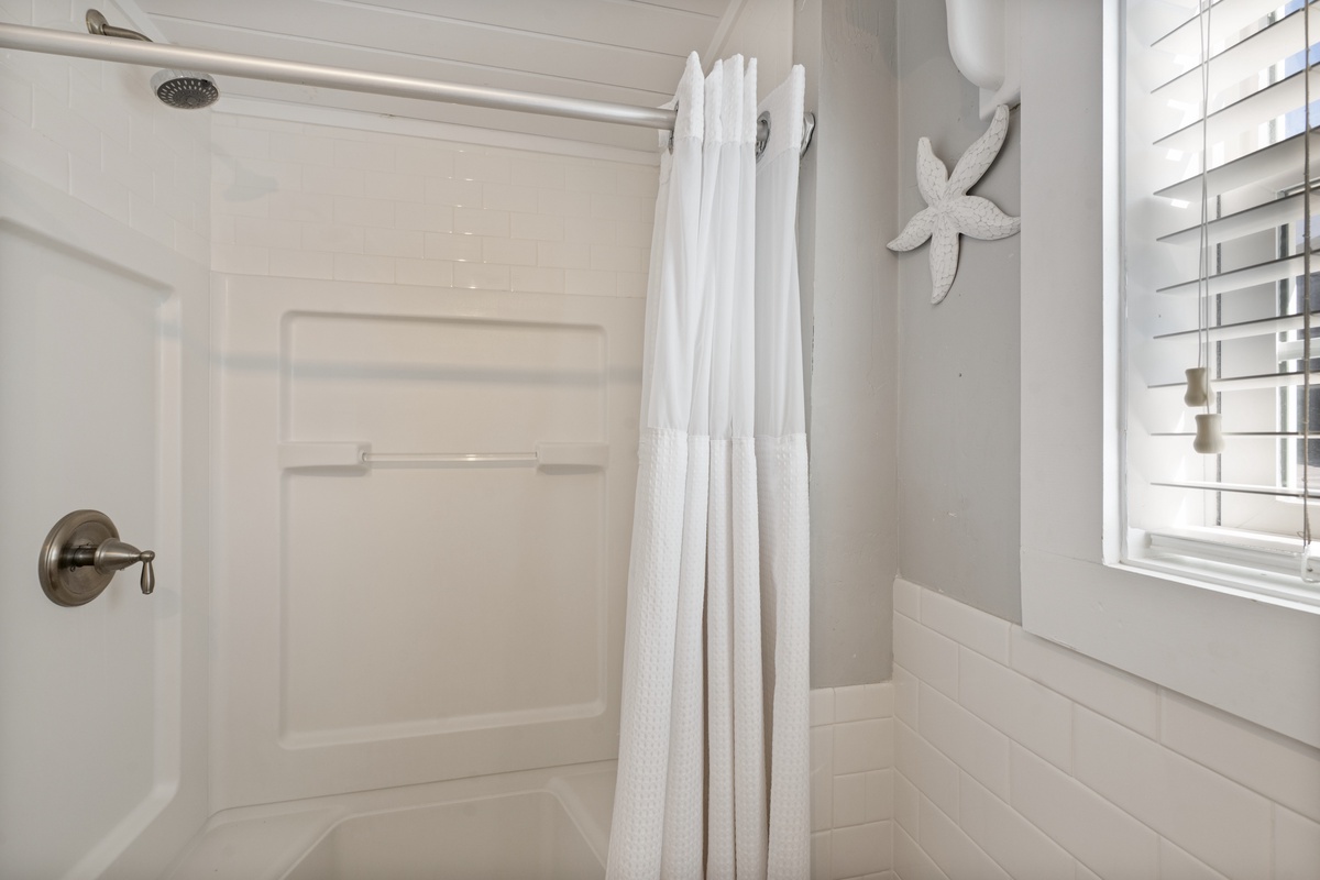 Refresh in your pristine white bathroom featuring a spacious tub-shower combo with coastal starfish charm and natural light streaming through plantation shutters.