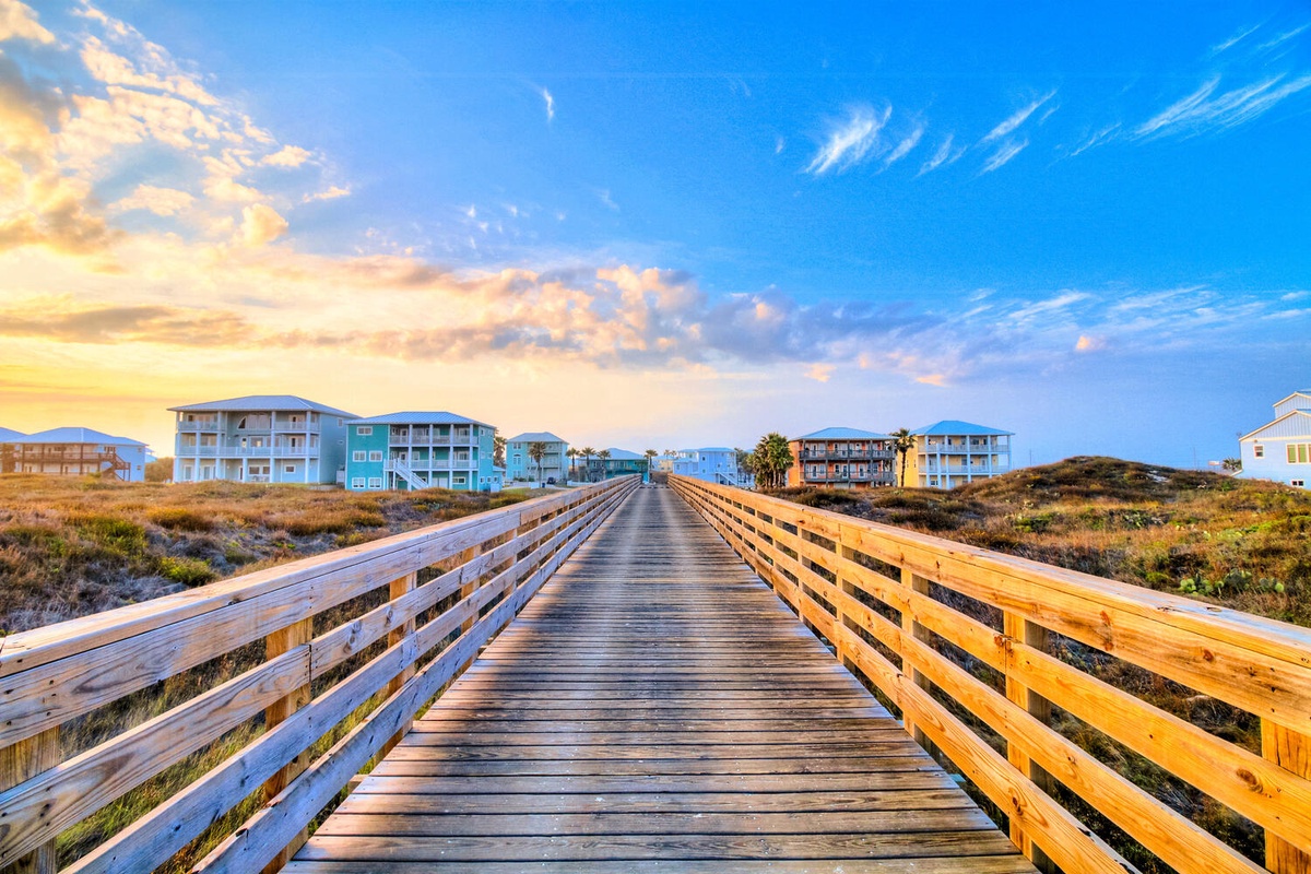 Wooden boardwalk leads through coastal dunes toward colorful beach houses beneath a stunning sunset sky.