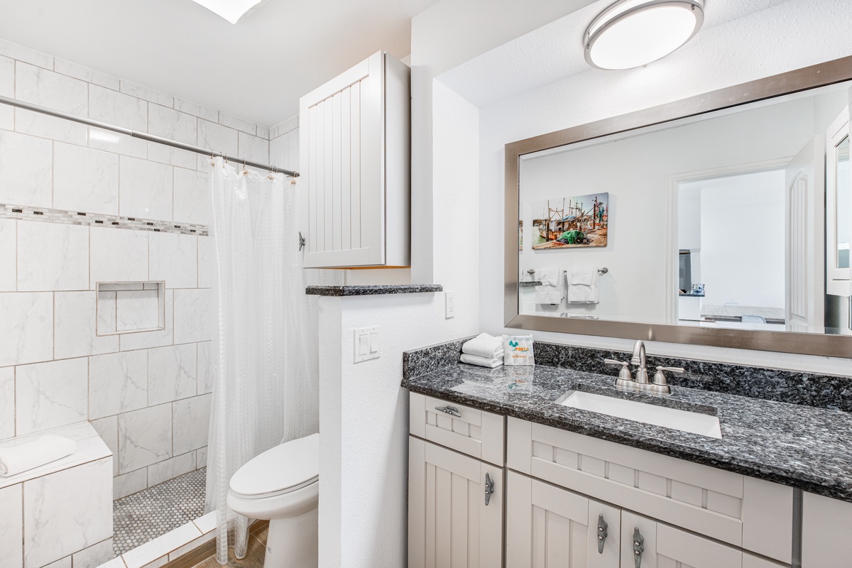 Your pristine bathroom features a spacious walk-in shower, elegant granite countertops, and coastal charm with fresh white cabinetry and soft lighting.