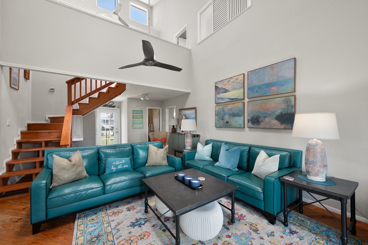 Sink into plush teal sofas beneath soaring ceilings where natural light floods the open living space. The warm wood staircase and coastal-inspired art create a welcoming retreat for your getaway.