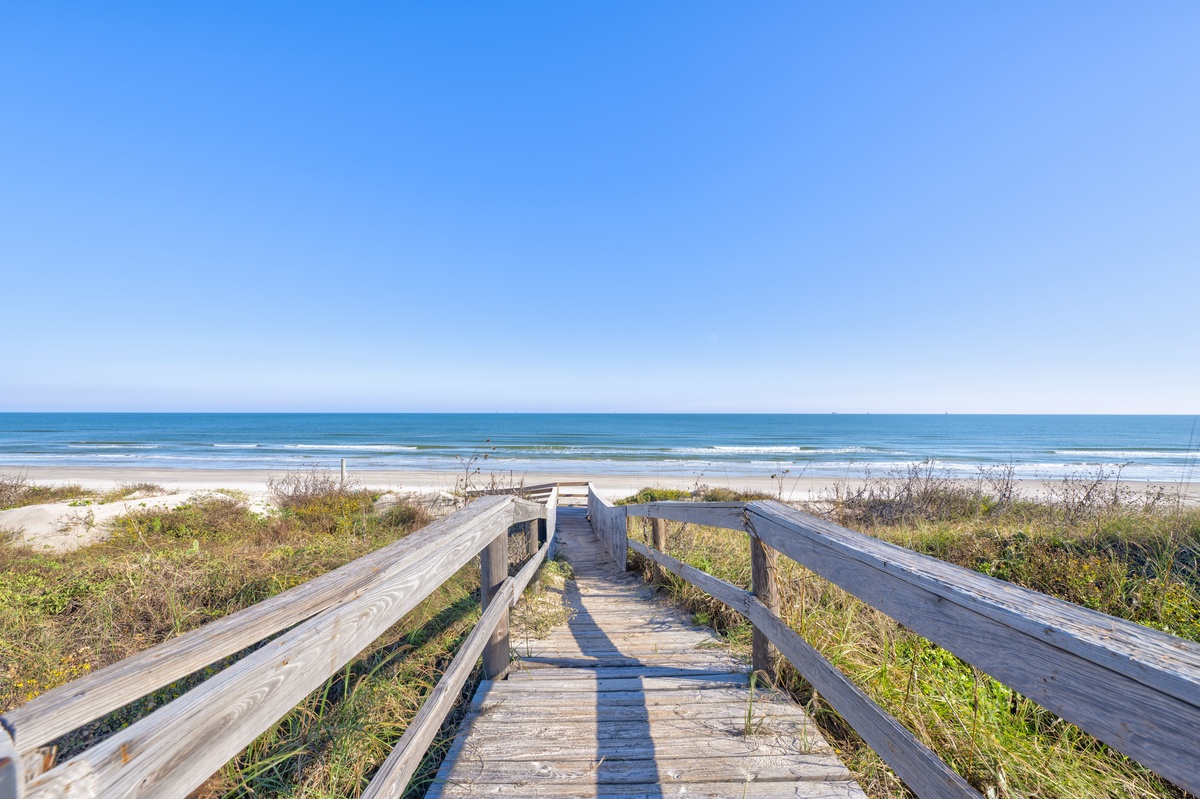 Wooden boardwalk leads through coastal dunes to pristine sandy beach and ocean waves under clear blue skies.