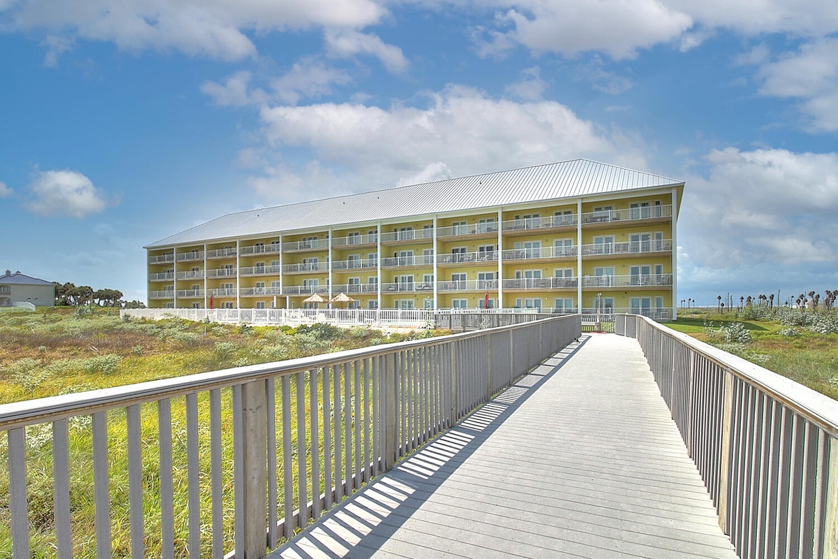 A modern beachfront resort with multiple balconied floors sits beyond coastal dunes, accessible via an elevated wooden boardwalk.