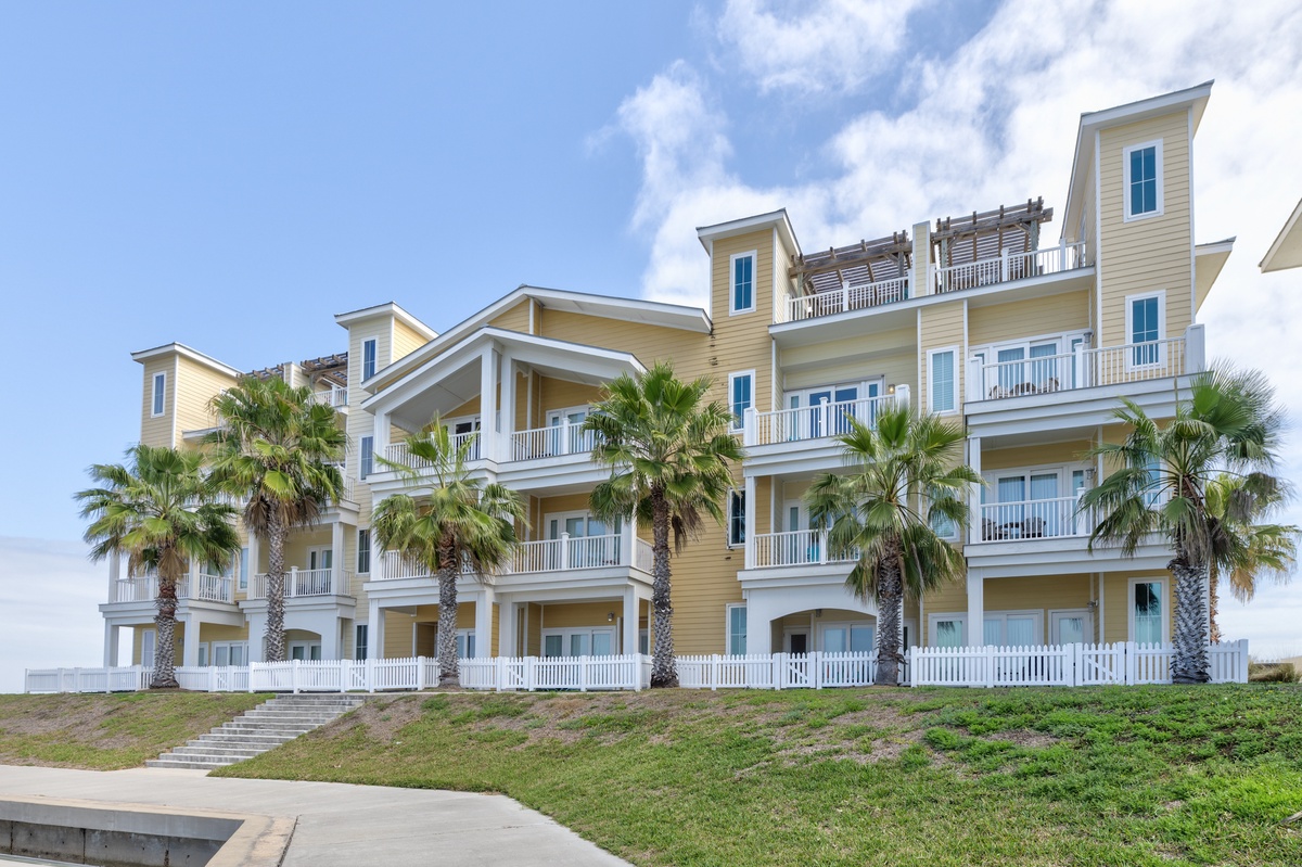 Coastal resort building featuring multiple balconies and tropical palm landscaping under sunny blue skies.