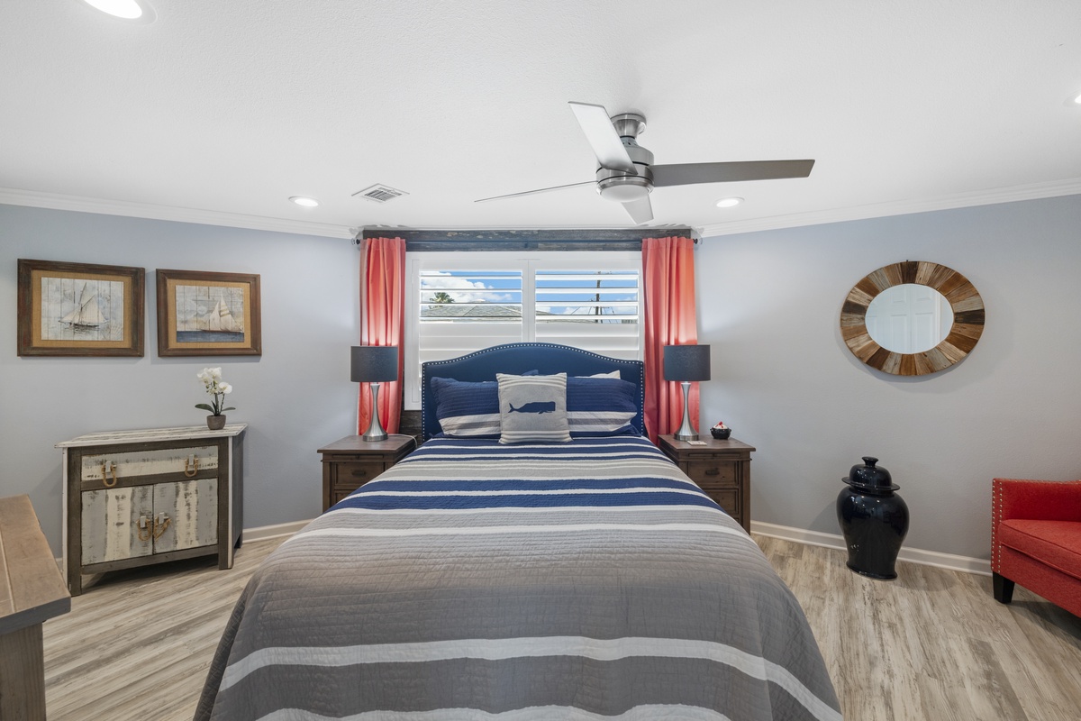 Your stylish bedroom features a plush blue headboard, crisp nautical-themed linens, and a ceiling fan for perfect comfort.