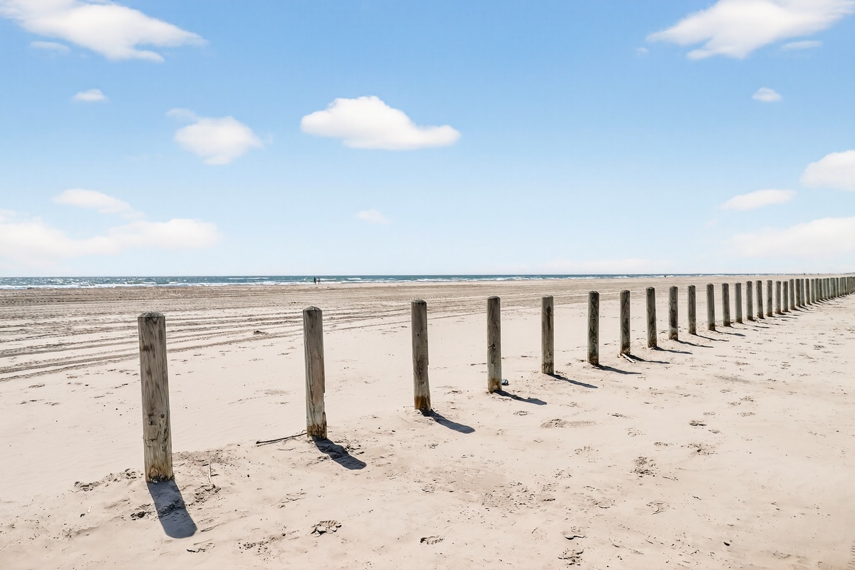 Pristine sandy beach with wooden posts stretching toward the ocean under a bright blue sky with fluffy white clouds.