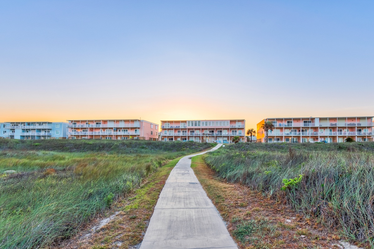 Peaceful walkway leads through coastal dunes to colorful beachfront buildings at golden hour.