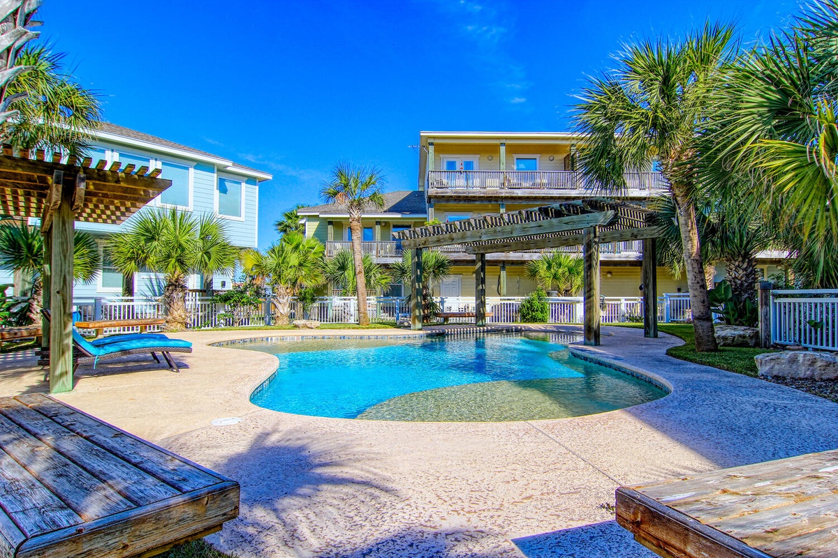 Beautiful resort-style pool area surrounded by tropical palms and colorful vacation homes under brilliant blue skies.