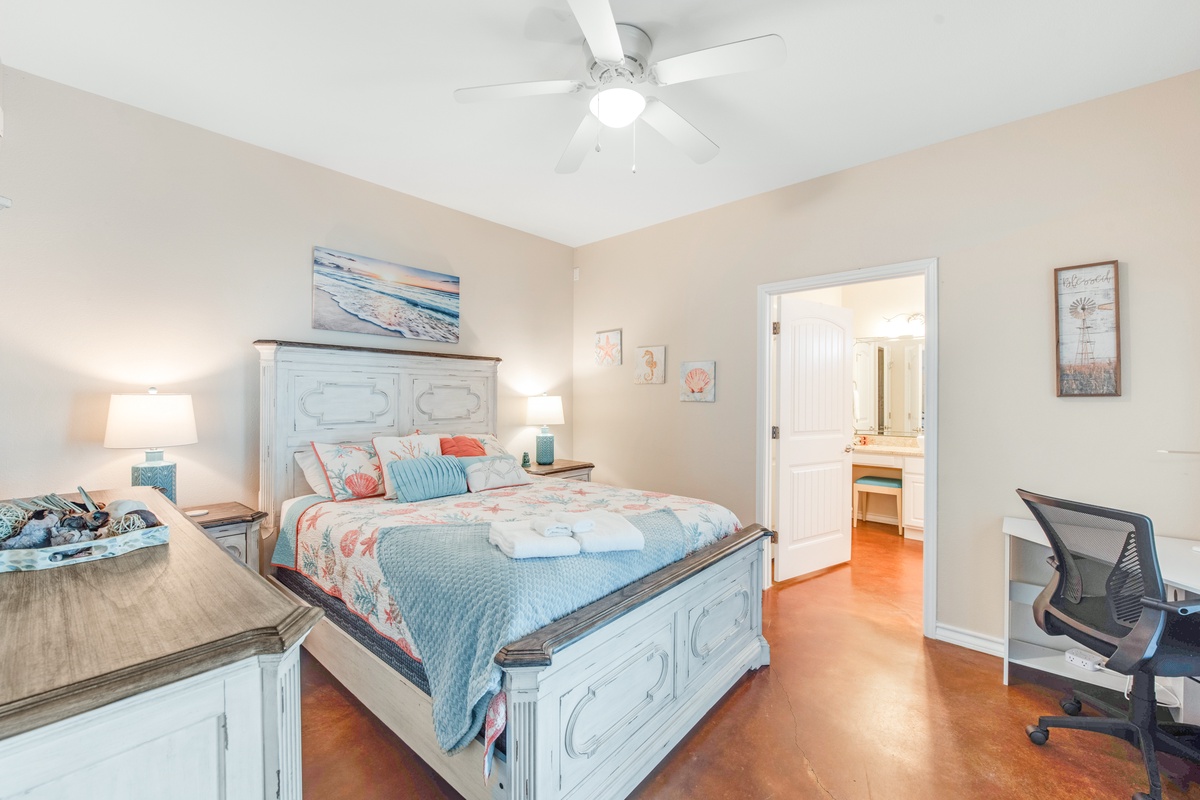 Unwind in your coastal-themed bedroom with plush bedding, gentle ceiling fan breeze, and convenient workspace for ultimate comfort.