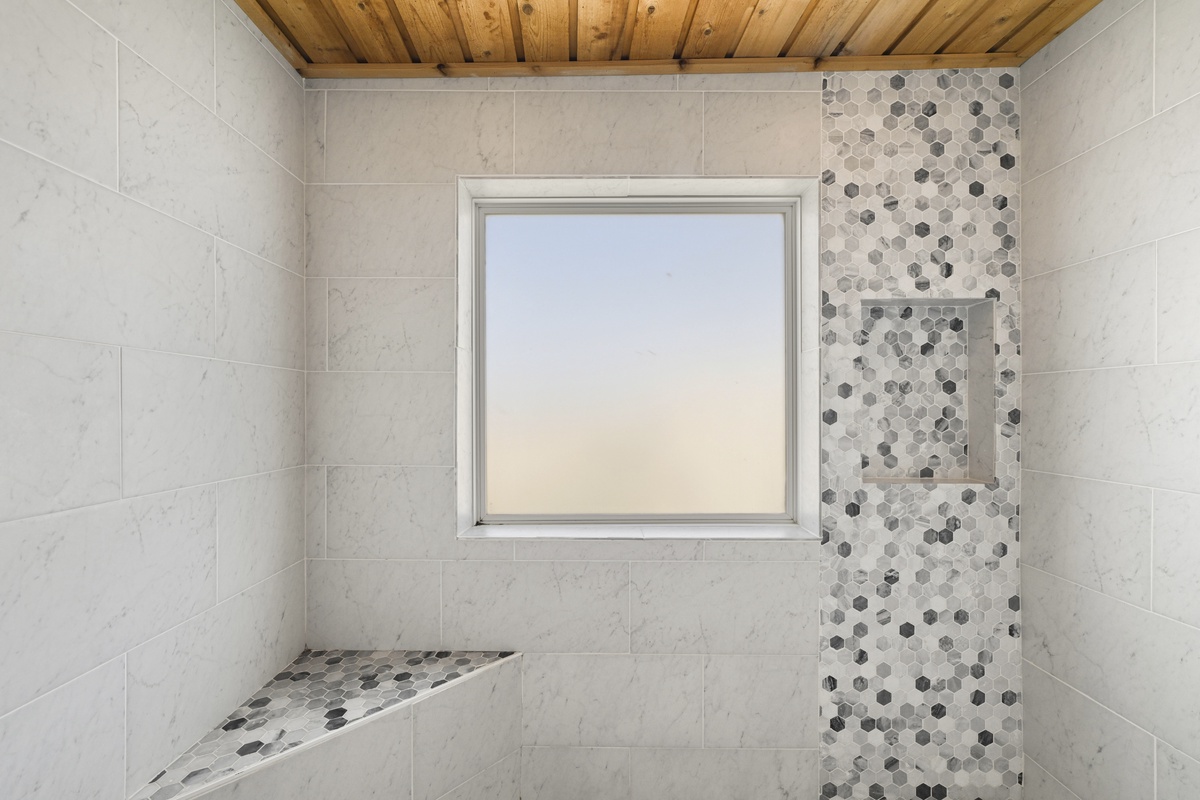 Your private sanctuary: a spacious walk-in shower with a built-in bench, elegant marble-style tile, decorative mosaic accents, natural light, and warm wood ceiling touches.