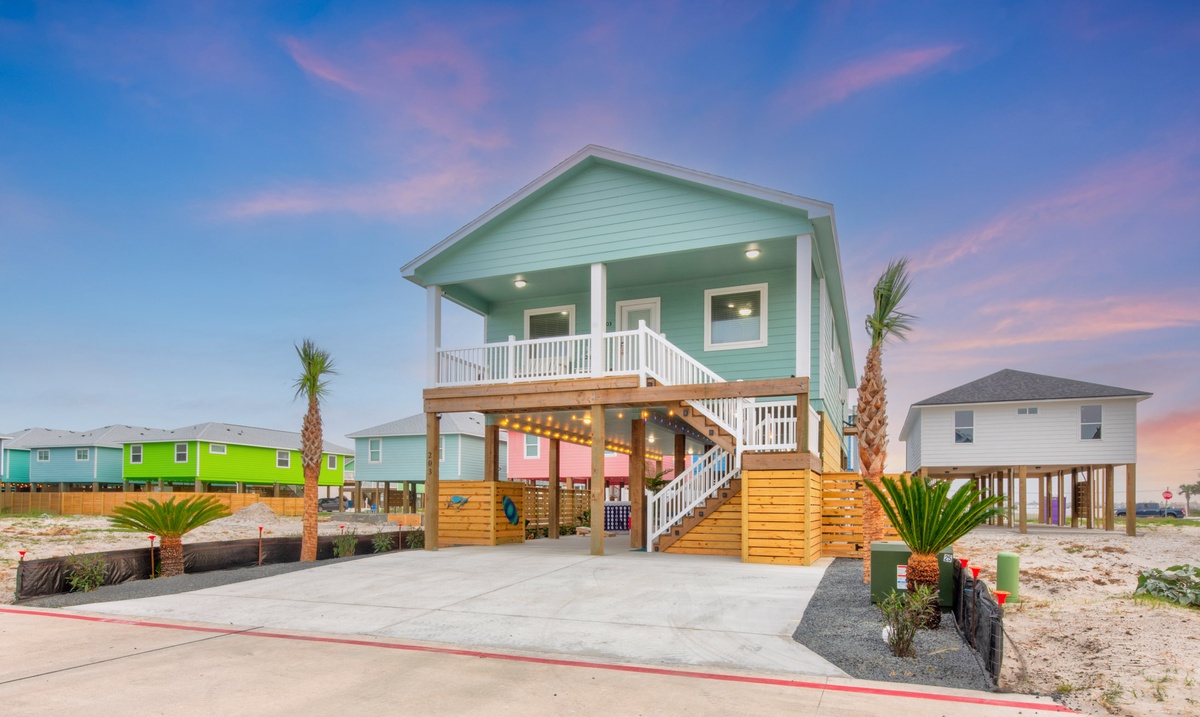 Charming coastal retreat with elevated design, featuring a welcoming covered porch and tropical landscaping beneath vibrant sunset skies.