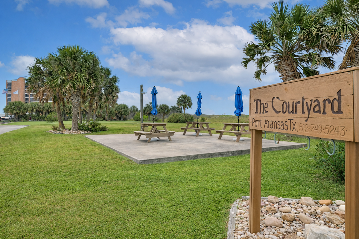 The Courtyard in Port Aransas offers a tranquil outdoor setting with picnic tables, palm trees, and green lawns under bright blue skies.