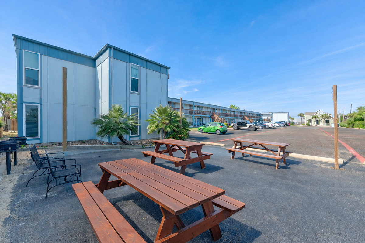 Modern beachside property featuring picnic tables and outdoor seating areas with tropical palm trees and clear blue skies.