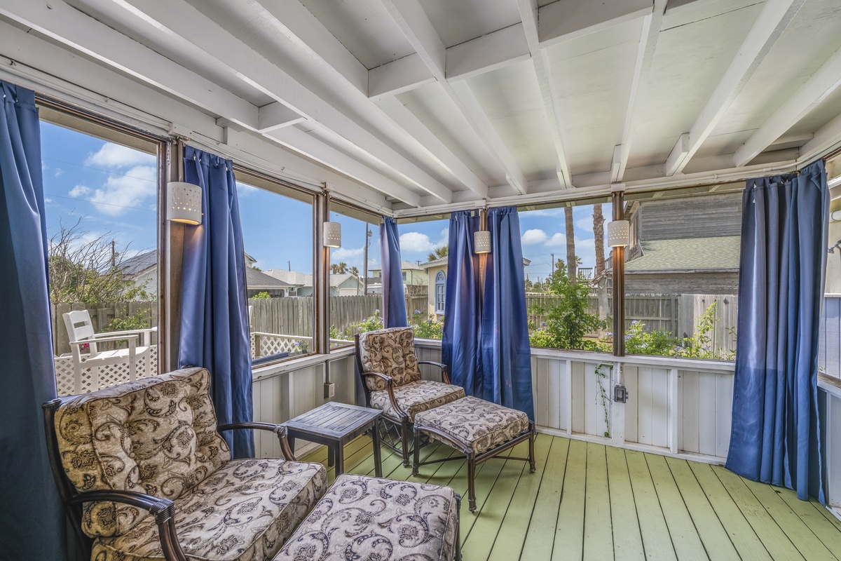 An enclosed sunroom overlooking the neighborhood, where you can unwind in cushioned chairs, enjoy natural light through large windows, and soak in peaceful garden views all around.