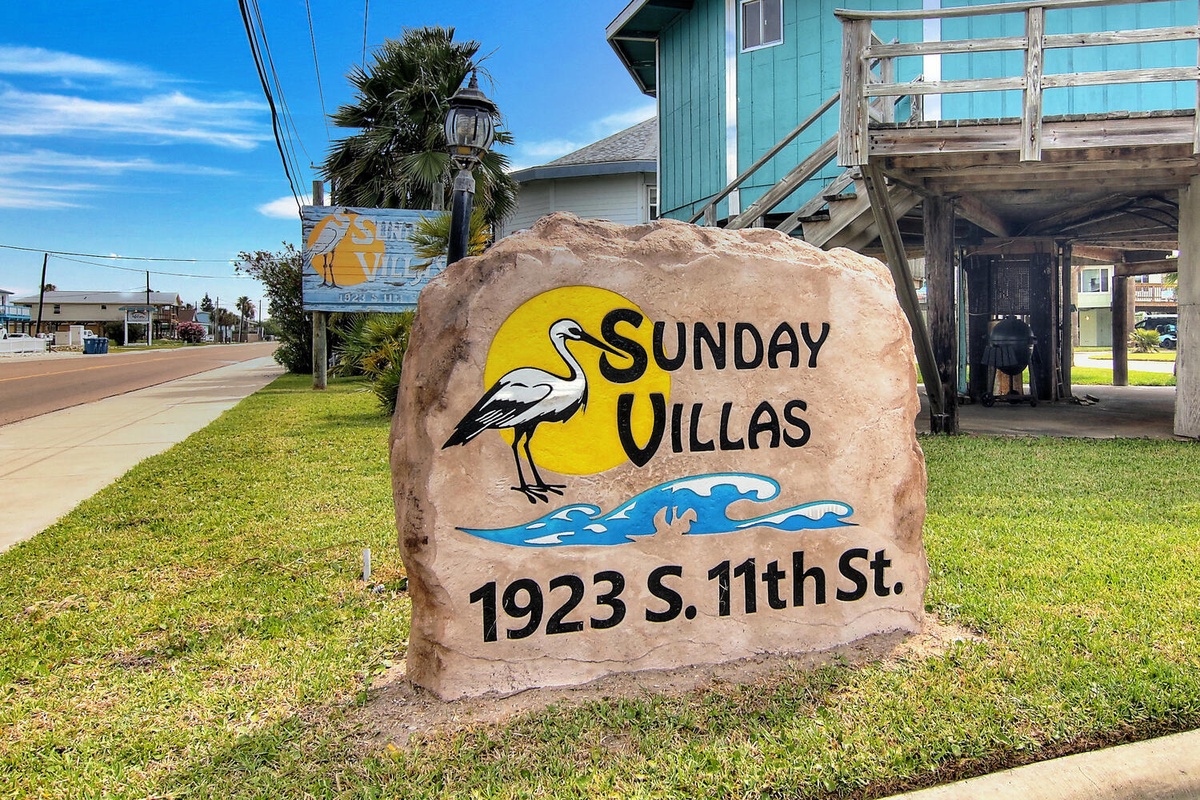 Welcome to Sunday Villas! This charming coastal property marker sits among palm trees and colorful beach houses.