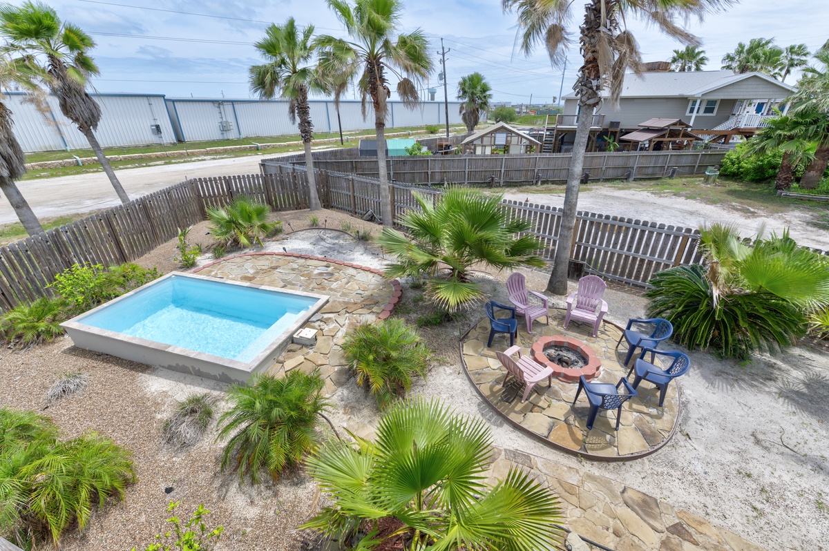 Aerial view of the rental property showcasing a private pool, fire pit area, tropical landscaping with palm trees, and fenced yard in a coastal neighborhood setting.