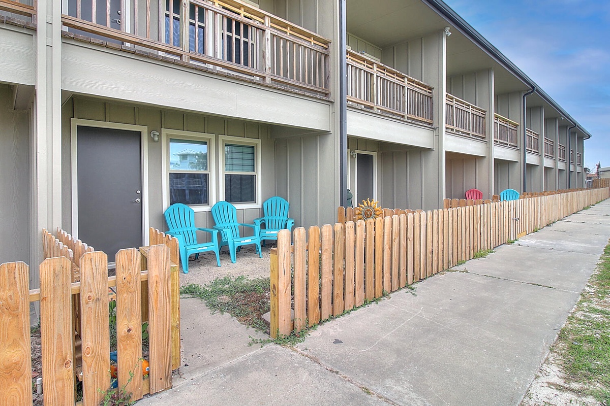 Multi-unit property building with individual patios featuring colorful outdoor seating behind wooden fencing.