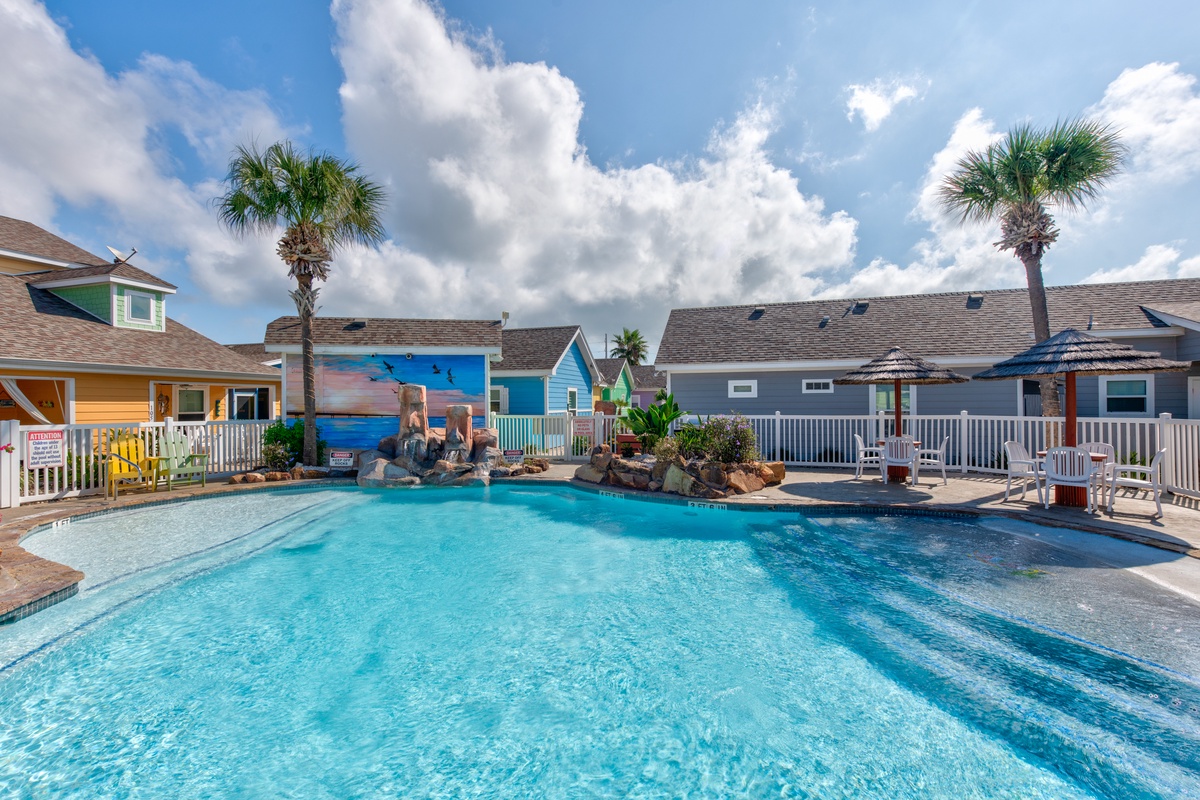 Tropical-themed resort pool surrounded by colorful beach houses and swaying palm trees under bright blue skies.