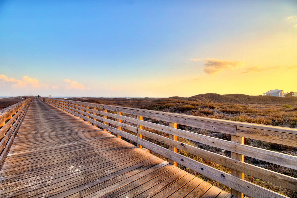 Wooden boardwalk stretches through coastal dunes toward golden sunset skies, offering scenic walking access to nearby natural areas.