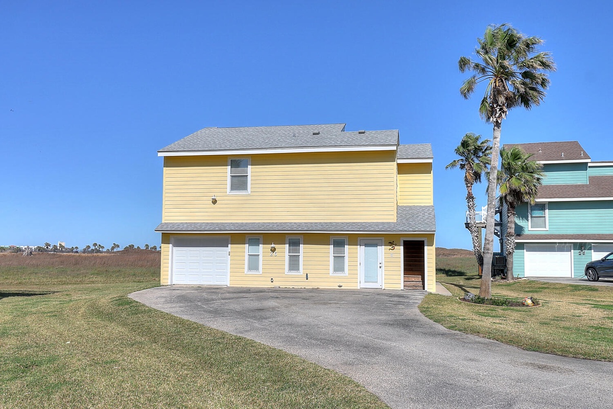 Two-story yellow beach house with palm trees and open grassy fields, featuring garage parking and coastal neighborhood charm.