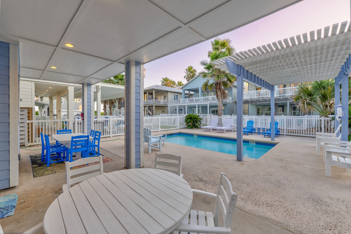 Relax at your covered poolside dining area with vibrant blue chairs and palm trees surrounding the sparkling pool.