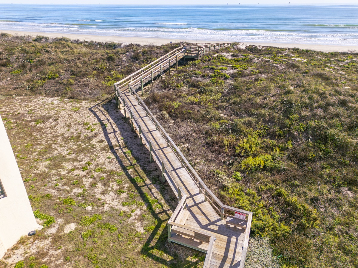 Elevated wooden boardwalk winds through coastal dunes to pristine sandy beach with rolling waves beyond.