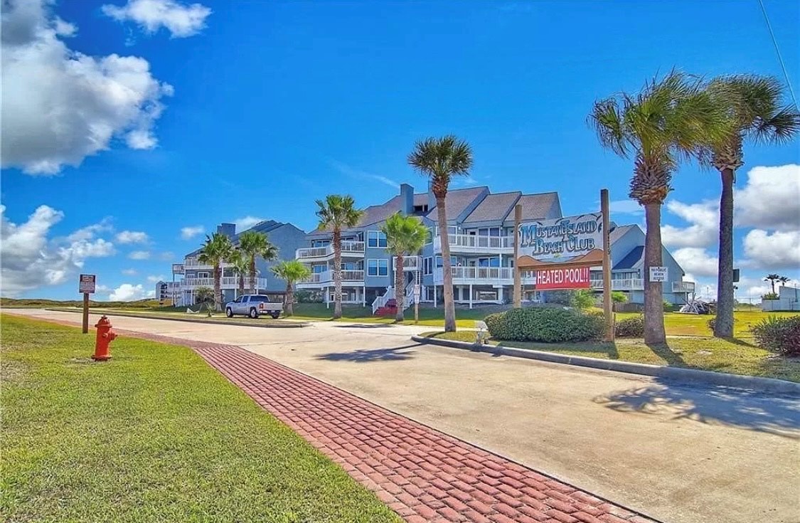 Mustang Island Beach Club welcomes guests with its distinctive signage and tropical palm-lined entrance on this sunny coastal day.