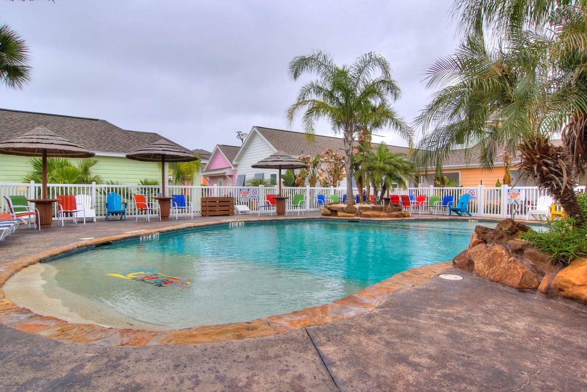 Colorful beachside resort with tropical pool area surrounded by palm trees and vibrant cottage-style buildings.