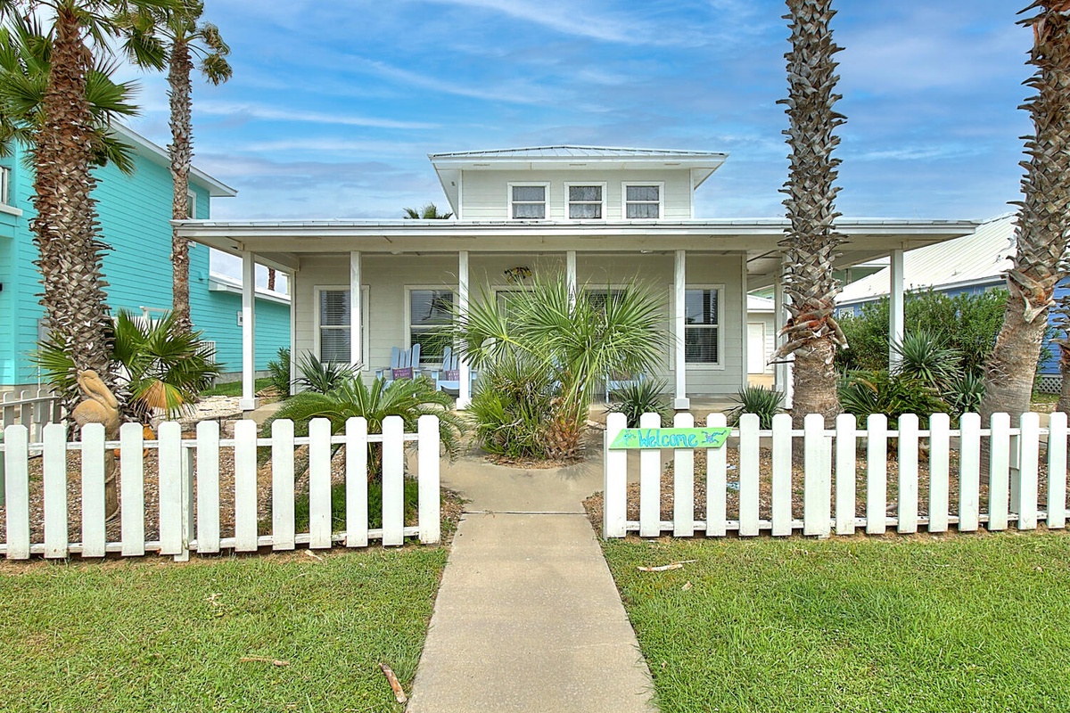 Charming coastal cottage with wraparound porch and tropical landscaping, featuring white picket fence and palm trees in beachside neighborhood.