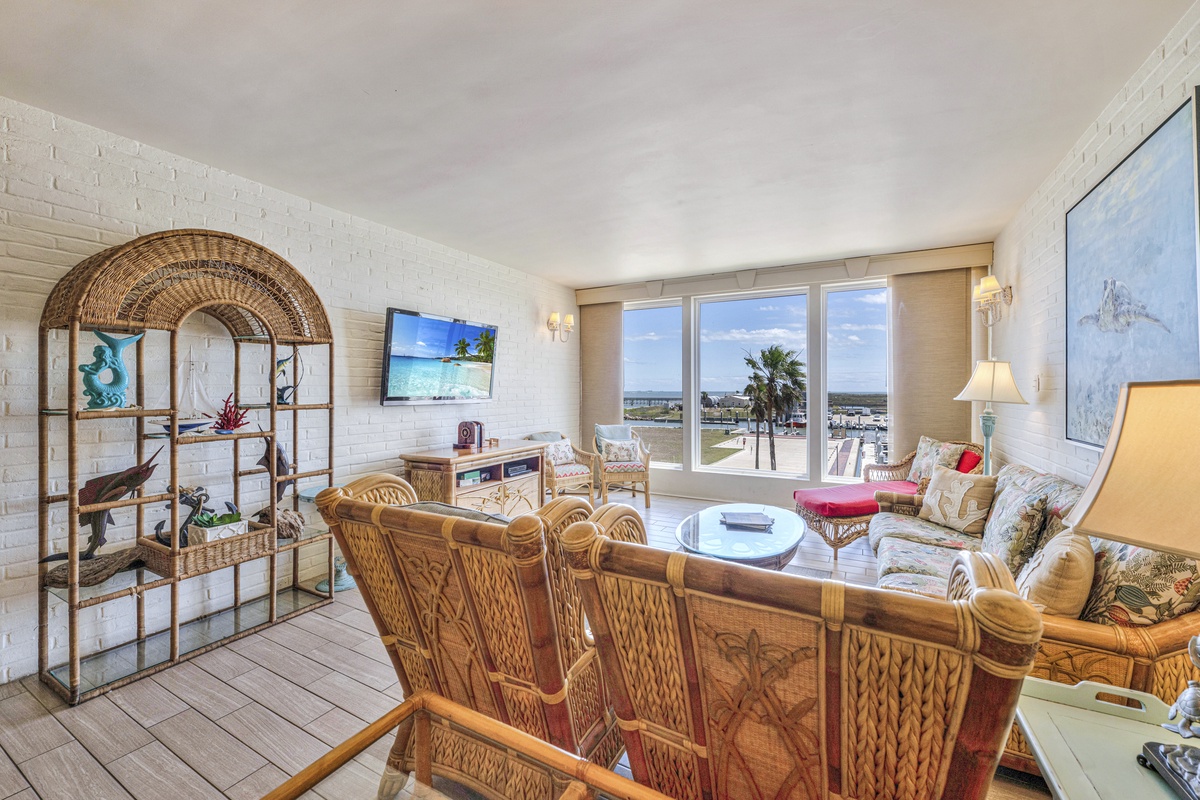 Your bright living room features tropical wicker furniture and stunning marina views through floor-to-ceiling windows, creating the perfect coastal retreat.