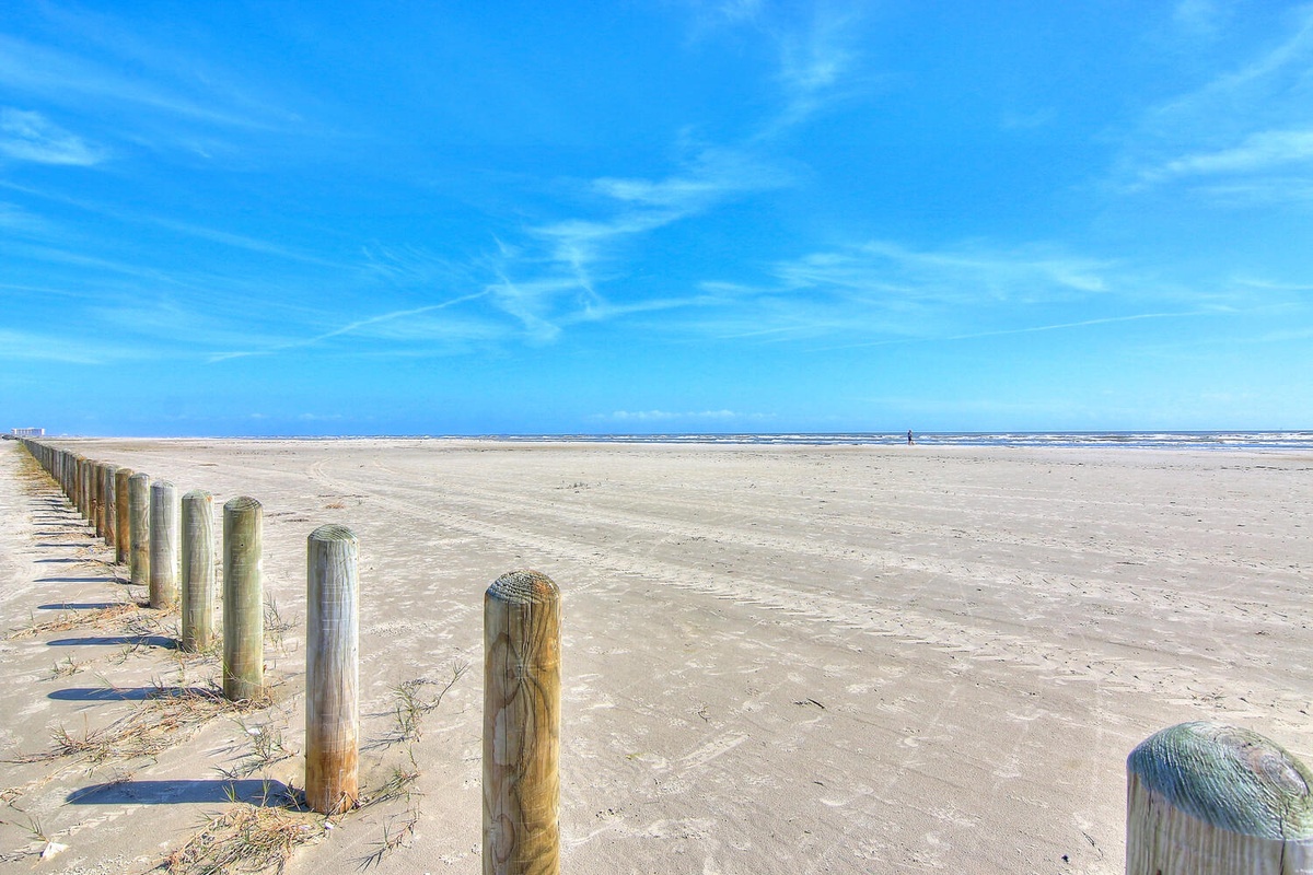 Pristine sandy beach stretches endlessly under brilliant blue skies, with weathered wooden posts marking the coastal boundary.