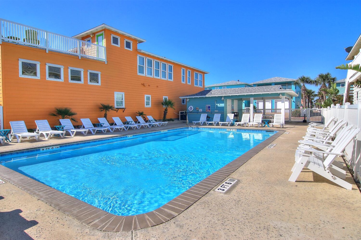 Sparkling pool with comfortable loungers surrounded by vibrant coastal buildings under endless blue skies.