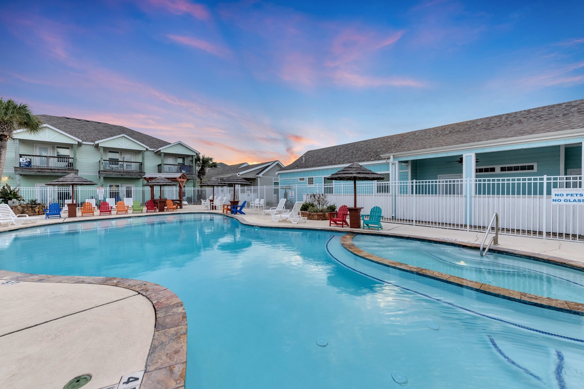 Resort pool area with colorful Adirondack chairs under a vibrant sunset sky, surrounded by coastal-style buildings.