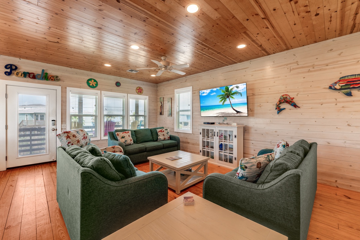 Gather with family in your cozy living room featuring warm wood walls, comfortable green seating, and tropical beach vibes on the big screen.