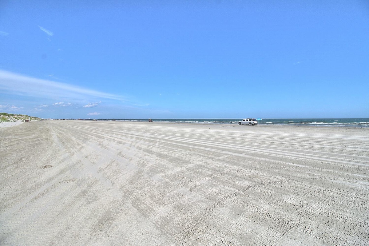 Expansive sandy beach stretches endlessly under brilliant blue skies, with gentle waves and scattered vehicles on the shore.