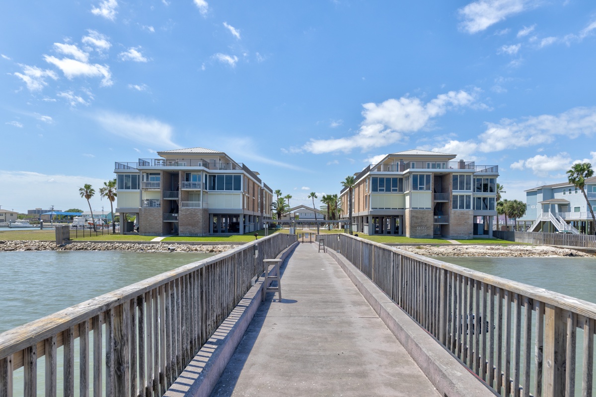 Modern waterfront buildings rise from the shoreline, connected by a private pier extending into calm coastal waters.