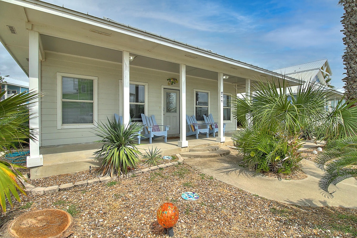 Charming coastal cottage featuring a welcoming covered porch with comfortable seating, surrounded by tropical landscaping.