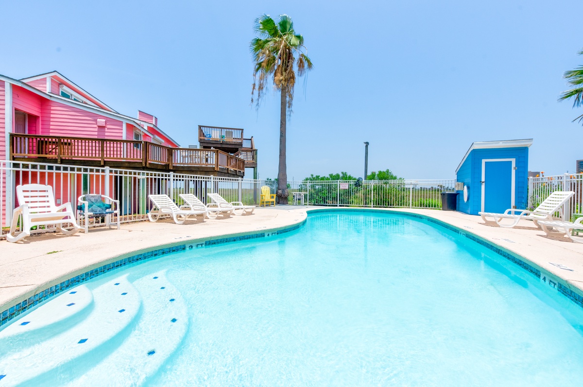 Vibrant coastal rental featuring a sparkling pool surrounded by colorful beach cottages and swaying palms under clear blue skies.