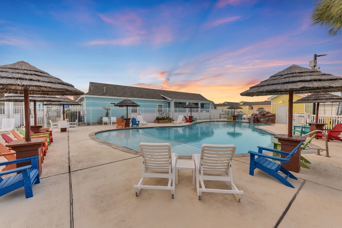 Resort-style pool area surrounded by thatched umbrellas and colorful Adirondack chairs under a stunning sunset sky.