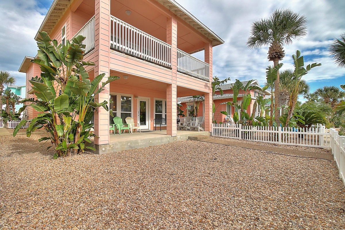 Bright coral-colored beach house with tropical landscaping, white picket fence, and upper balcony in a coastal community setting.