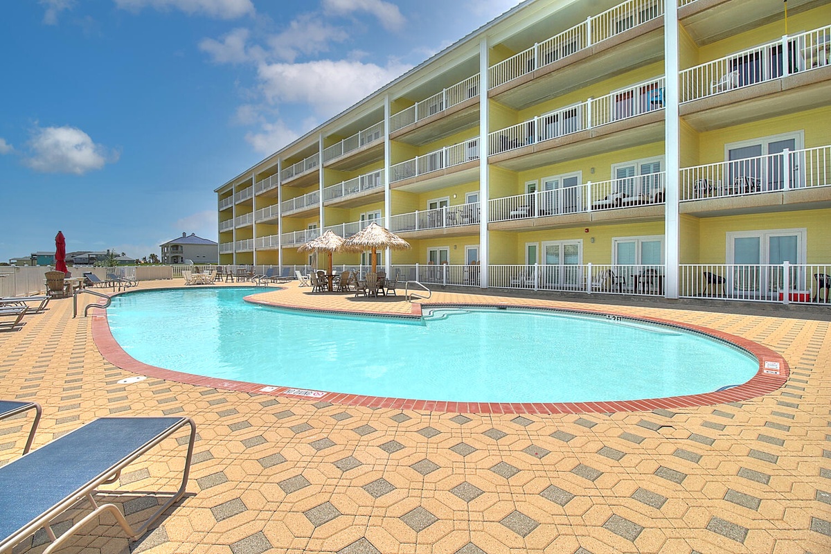 Bright yellow resort building with curved outdoor swimming pool surrounded by brick pavers and multiple balconied guest rooms.