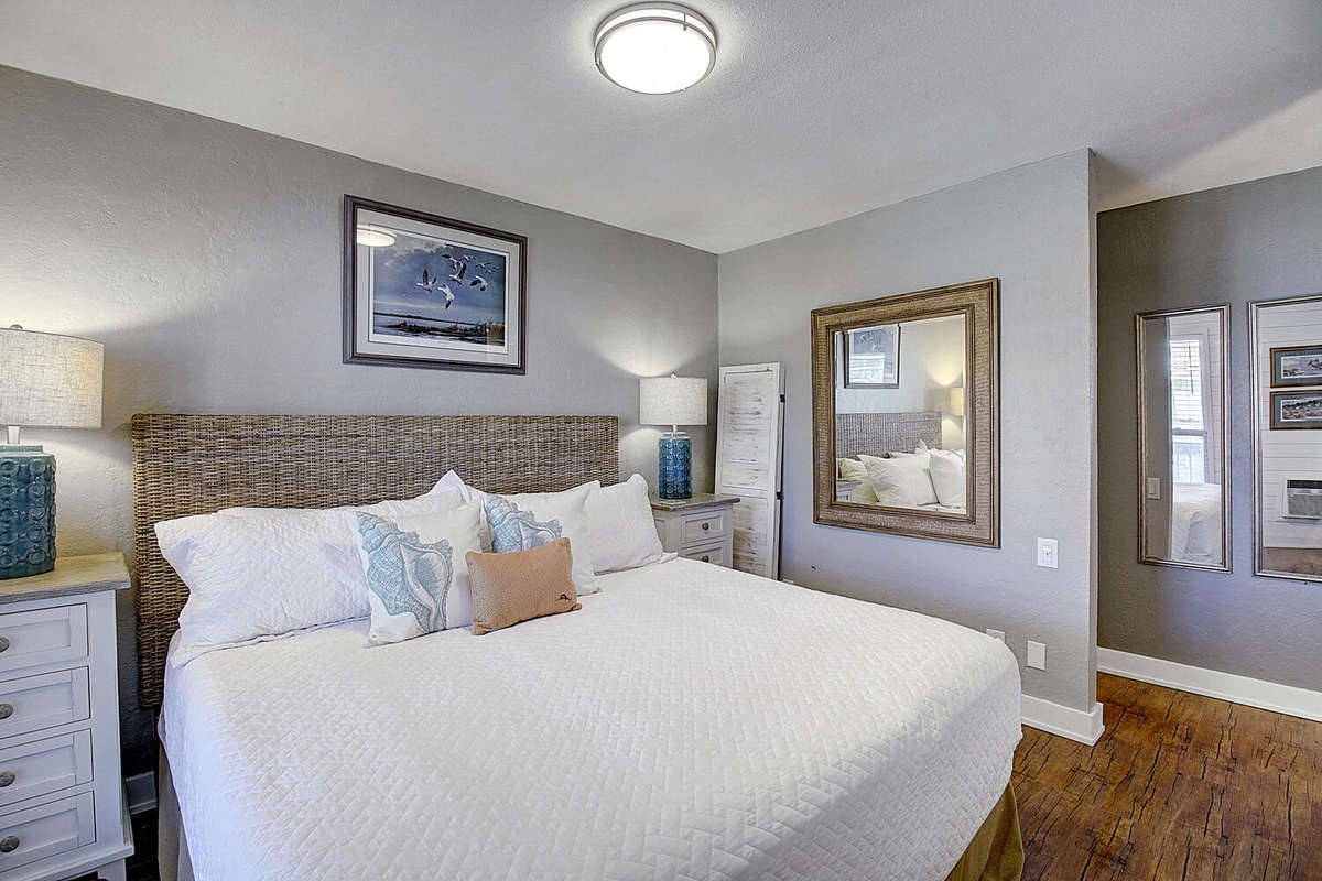 Drift off in your cozy bedroom featuring a plush bed, soft linens, and coastal charm with warm wood floors underfoot.