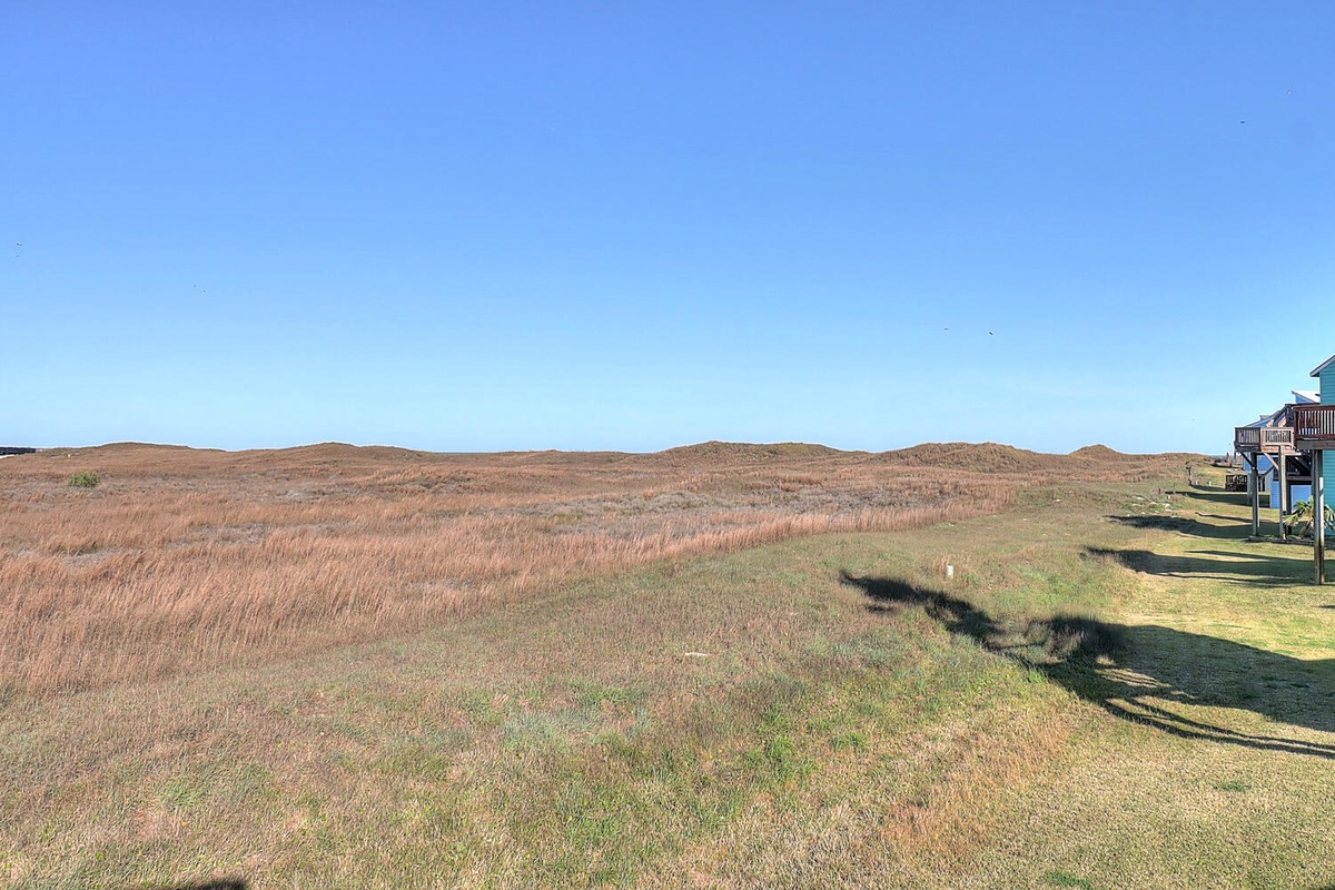 Expansive coastal marshland stretches toward rolling hills under clear blue skies, with the property's elevated deck providing scenic overlooks of this pristine natural setting.