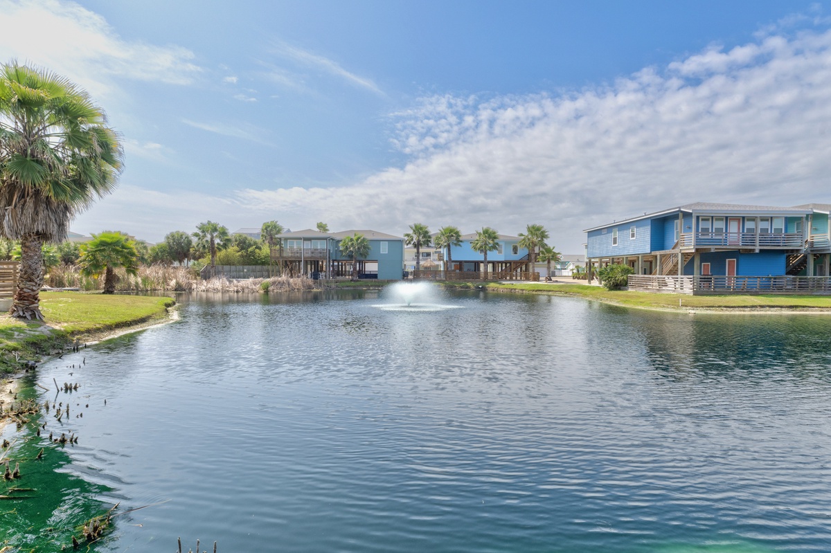 This waterfront community features colorful beach homes along a serene pond with a fountain centerpiece, surrounded by palm trees and coastal vegetation under open skies.