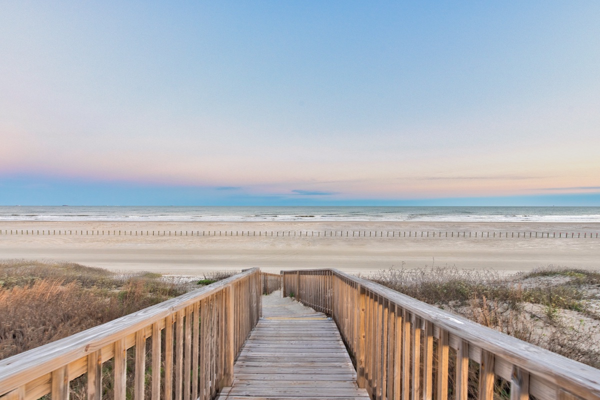 Wooden walkway leads directly to pristine beach with gentle waves and endless ocean horizon at sunset.